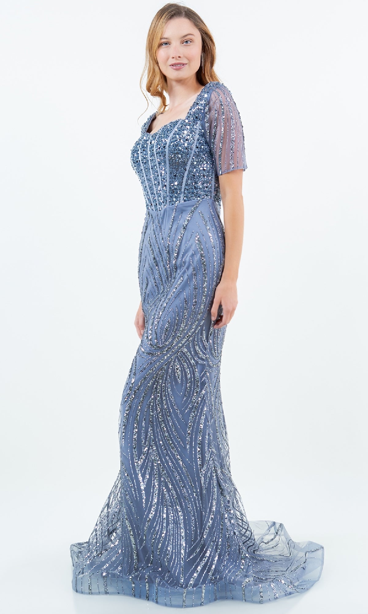 A woman poses against a white background in the Cinderella Couture 8293M, a long sequin formal gown with light blue coloring, floor-length mermaid silhouette, short sheer sleeves, and silver sequin patterns.